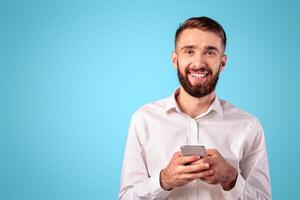 Cheerful millennial guy with smartphone browsing web, playing game, shopping online on blue studio background. Positive Caucasian man with modern gadget looking at camera and smiling photo