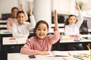 Concept of School Reopen, Back To Normal Lifestyle. Portrait of excited little schoolgirl raising hand up in the air to answer a question with all her classmates in blurred background, selective focus photo