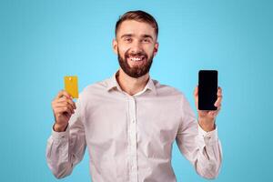 Happy young guy showing credit card and smartphone with empty screen on blue studio background, mockup for mobile app or website design. Satisfied client recommending online bank services photo