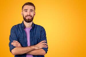 Joyful young guy with happy face posing with crossed arms over orange studio background. Portrait of positive bearded man in casual outfit looking at camera and smiling photo
