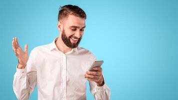 Smiling bearded guy using smartphone, surfing internet, communicating online over blue studio background. Happy millennial man working or studying remotely using mobile device photo