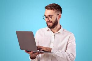 Successful young guy in formal wear using laptop computer over blue studio background. Cheerful businessman having online job, participating in web conference, communicating via internet photo