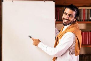 Remote modern education. Indian teacher man teaching online pointing to blackboard explaining English grammar rules standing indoors, smiling to camera. Guy teaches language distantly photo