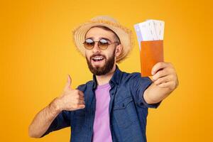 Overseas trip. Millennial guy holding international passport with airplane tickets and showing thumb up gesture over orange studio background. Cool tourist with documents ready to travel abroad photo