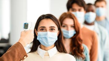 Woman Doing Temperature Screening Measuring Fever For Diverse People Standing In Queue, Using Infrared Contactless Themometer Indoors. Coronavirus, Covid-19 Pandemic Outbreak Concept photo