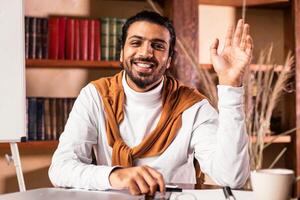 Joyful Indian Teacher Man Waving Hello To Camera Posing Sitting In Classroom With Blackboard Having Online Class Via Call. Distant Learning And Teaching, Virtual Lecture And Webinar Concept photo