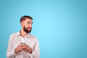 Happy young businessman with mobile phone looking at empty space on blue studio background. Smiling Caucasian guy using modern device for online work or studies, surfing web, checking email photo