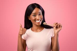 Pretty black lady with beautiful smile holding wooden toothbrush and showing thumb up gesture on pink studio background. Happy African American woman taking care of her teeth health photo