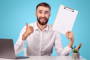 Happy Caucasian guy holding resume and showing thumb up gesture at his desk, blue studio background. HR manager finding perfect worker for business team. Successful employment and headhunting concept photo