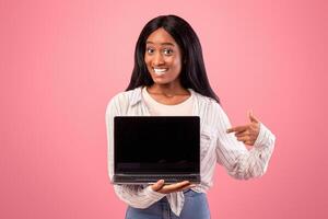 Happy black woman pointing at laptop with empty screen and smiling on pink studio background. Positive African American female with portable computer, mockup for design. Online work, education photo