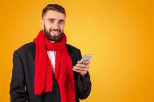 Handsome businessman in elegant jacket and red scarf using mobile phone over orange studio background. Positive young guy searching web, watching news or participating in online conference photo