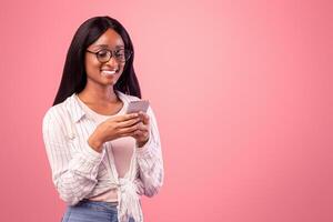 Online life. Smart black woman in glasses using cellphone for remote work or studies on pink studio background. Intelligent African American business lady having web conference on mobile device photo