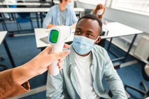 Teacher using digital medical contactless infrared electronic thermometer, measuring and checking body temperature, screening black student in protective mask, preventing contagious diseases spread photo