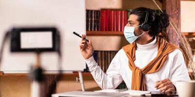 Virtual Education. Indian Male Teacher In Face Mask Teaching Online Recording Lecture For Remote Students Explaining English Grammar, Pointing To Blackboard Sitting Indoors. Selective Focus photo