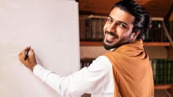 Cheerful Arab Teacher Guy Writing English Rules On Blackboard Having Online Lecture Indoor, Smiling To Camera. E-Learning And E-Teaching, Modern Virtual Education Concept photo