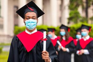 Black guy student in graduation costume showing diploma at camera, posing over international group of students at university campus, wearing protective face mask, panorama with copy space photo