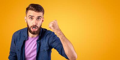 Portrait of young angry guy shaking clenched fist at camera and shouting, showing his protest over orange studio background. Furious millennial man threatening someone, expressing negativity photo