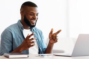 Online Communication. Happy African Man Using Laptop For Call, Enjoying Web Conference With Friends Or Family, Cheerful Black Guy Sitting At Desk With Computer, Taking And Gesturing At Camera, photo