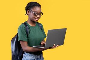 Cheerful Black Student Woman Using Laptop Standing Over Yellow Studio Background, Side View. Remote Education, E-Learning And Online Study Concept. Panorama With Copy Space photo