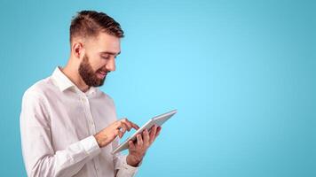 Confident young guy in office wear using tablet computer over blue studio background. Focused freelancer working or studying remotely, having web conference, communicating online photo