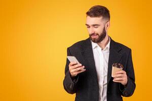 Positive young businessman checking email or browsing web on his cellphone, drinking takeaway coffee on orange studio background. Happy guy in formal wear using internet on mobile device photo