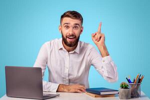 Millennial Caucasian office worker having creative idea, gesturing eureka at workplace on blue studio background. Young man finding solution to problem, sitting at his desk with laptop photo