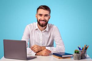 Successful young office worker sitting at desk with laptop computer and smiling on blue studio background. Portrait of happy millennial businessman in formal wear at workplace photo
