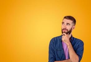 Dreamy hipster guy thinking over something, looking at empty space on orange studio background. Pensive young man making choice or decision, being thoughtful, having doubts photo