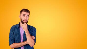 Portrait of attractive young guy thinking and touching his chin over orange studio background, copy space. Pensive millennial man having doubts, making decision or choice, dreaming about something photo