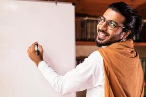 Distance Education. Happy Indian Teacher Man Teaching Remote Students Online Pointing To Blackboard With English Language Rules Standing Smiling To Camera At Home. Knowledge, E-Teaching And Learning photo
