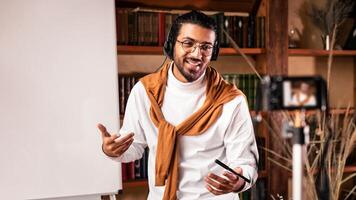 Indian Tutor Guy Making Lecture For Educational Blog Explaining English Grammar Rules To Camera Standing Near Blackboard At Home. Modern Teacher, Remote Education And E-Teaching Concept photo