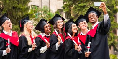 Cheerful multiracial group of students in graduation dresses taking selfie on smartphone, holding diplomas and posing over university building, enjoying and celebrating graduation photo
