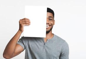 Hi friend. Smiling african-american man covering half of his face with clean sheet of paper, copy space photo