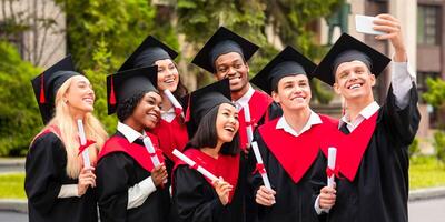 Positive multiracial group of students in graduation dresses taking selfie on smartphone, holding diplomas and posing over university building, enjoying and celebrating graduation photo