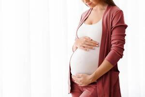 Expecting A Baby. Portrait Of Happy Young Pregnant Lady Tenderly Touching Belly Near Window At Home, Beautiful Millennial Awaiting Mother Embracing Her Tummy And Smiling, Enjoying Pregnancy Time photo