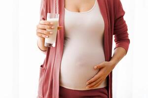 Dairy Products During Pregnancy. Closeup Shot Of Pregnant Lady Holding Glass Of Milk, Unrecognizable Expectant Mother Standing Near Window At Home And Embracing Her Belly, Cropped Image, Free Space photo