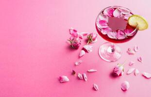 Cocktail bar background. Rose beverage with vodka, pineapple juice and strawberry champagne served with lemon slice and rose petals at pink backdrop, copy space photo