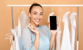 Online Sales And Mobile Application. Smiling lady showing smartphone with black empty screen for mock up template and holding hangers with clothes, standing and posing at studio near fashion rack photo