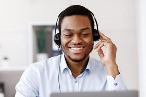Closeup of black guy worker with headset using laptop, cheerfully talking to client or business partner, smiling at computer screen, african american man having business training, copy space photo