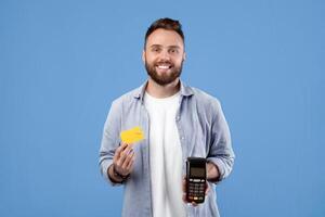 Smiling millennial guy holding payment terminal and credit card, looking at camera on blue studio background. Cool young man using electronic currency, preferring contactless shopping photo