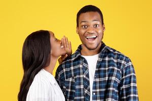 Amazing information, offered and secret for friend. Shocked millennial african american guy with open mouth listening to woman and look at camera isolated on yellow background, studio shot, copy space photo