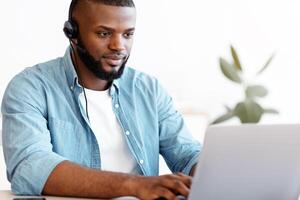 Portrait of african american man in headset working on laptop in office, millennial black male customer support hotline manager sitting at desk and looking at computer screen, free space photo