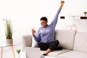 Weekend Leisure. Excited blackwoman listening to music in wireless earbuds, sitting on couch and using smartphone gadget, dancing and raising hand up in the air, enjoying favorite song in living room photo