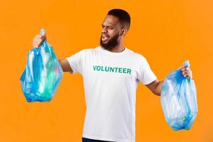 Displeased African American Volunteer Guy Holding Two Plastic Bags Full Of Garbage Litter And Bottles Posing In Studio Over Yellow Background. Ecology Problems And Pollution, Sort Your Waste Concept photo