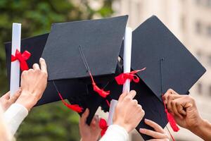 Graduation hats and diplomas raised up in students hands over university or college building, closeup. Cropped of multiracial group of students raising up hands with caps and diploma scrolls photo
