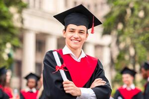 Cheerful guy in graduation costume showing his diploma and smiling at camera, happy male student posing over international group of students at university campus, copy space photo