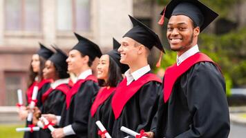 Diverse international students with diplomas attending graduation ceremony, happy multiracial group of students in graduation caps and robes cheerfully standing in a row at university campus photo