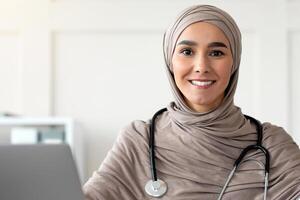 Beautiful muslim woman in hijab doctor with stethoscope posing in hospital, sitting at worktable with laptop, medical folder, smiling at camera, copy space. Doctor's office, cabinet interior concept photo
