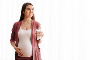 Happy Pregnant Lady Enjoying Healthy Calcium Drink, Holding Glass With Milk While Standing Near Window At Home, Young Smiling Expectant Lady Tenderly Embracing Belly And Looking Away, Copy Space photo