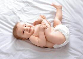 Beautiful caucasian baby laying on bed, playing with feet and smiling at camera, top view, copy space. Adorable toddler in diaper having fun alone, lying on white, laughing. Toddler, baby, infant photo